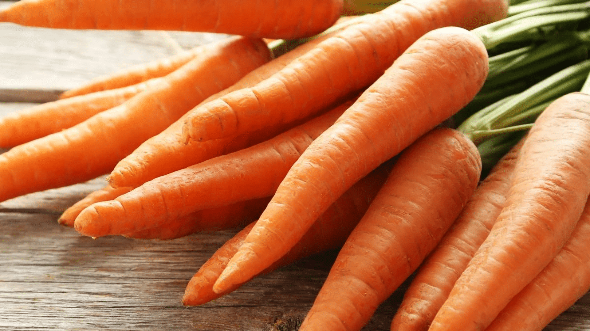 fresh orange carrots with green tops attached, stacked together on a wooden surface with natural texture visible