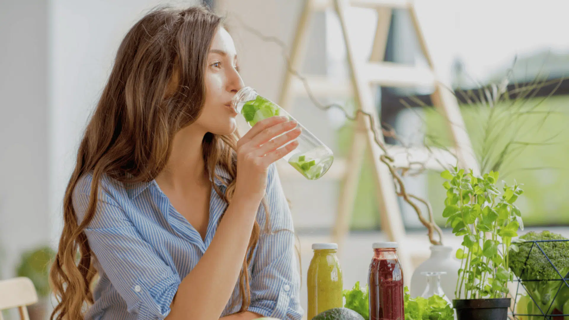woman drinking a green detox drink from a glass bottle while sitting at a table with fresh juices and herbs nearby