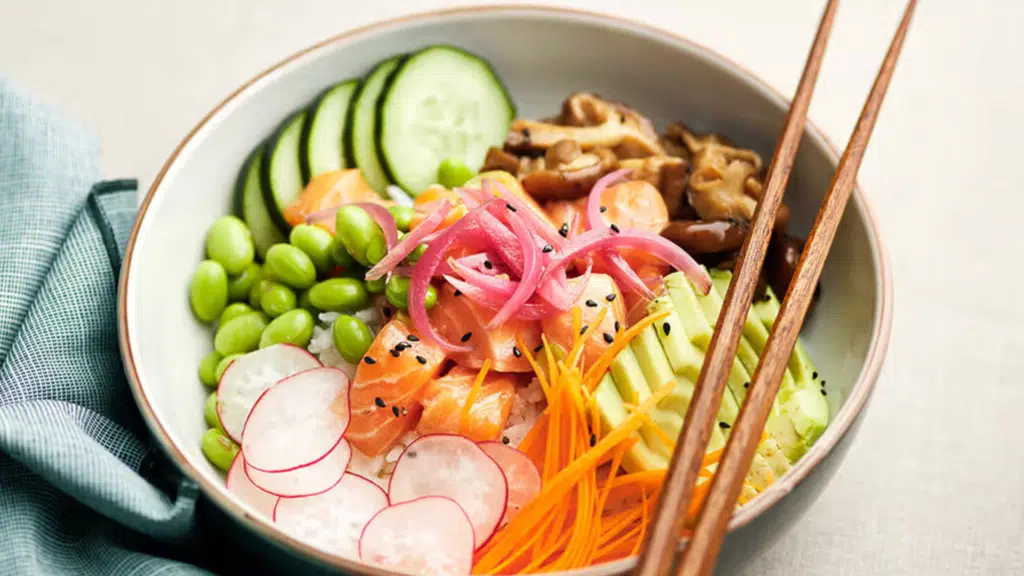 salmon poke bowl with rice edamame cucumber avocado radish pickled onions carrots and sesame seeds with chopsticks