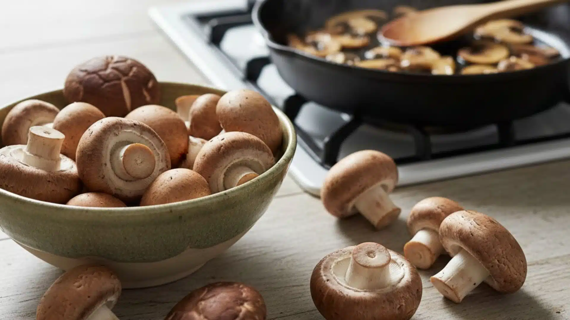 Fresh mushrooms in a bowl and scattered on a table, with a pan of sautéed mushrooms behind.