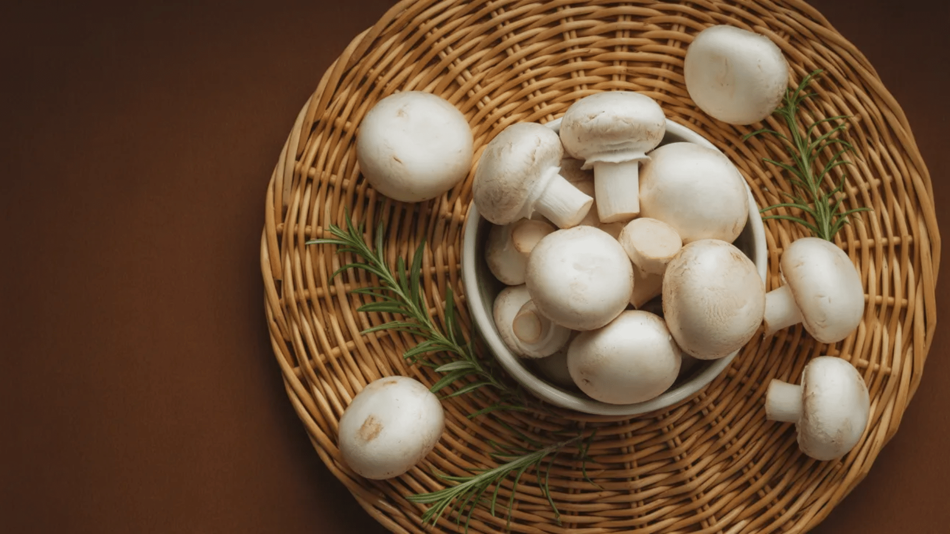 a bowl of fresh white button mushrooms and rosemary sprigs rests on a woven wicker tray against a warm brown backdrop