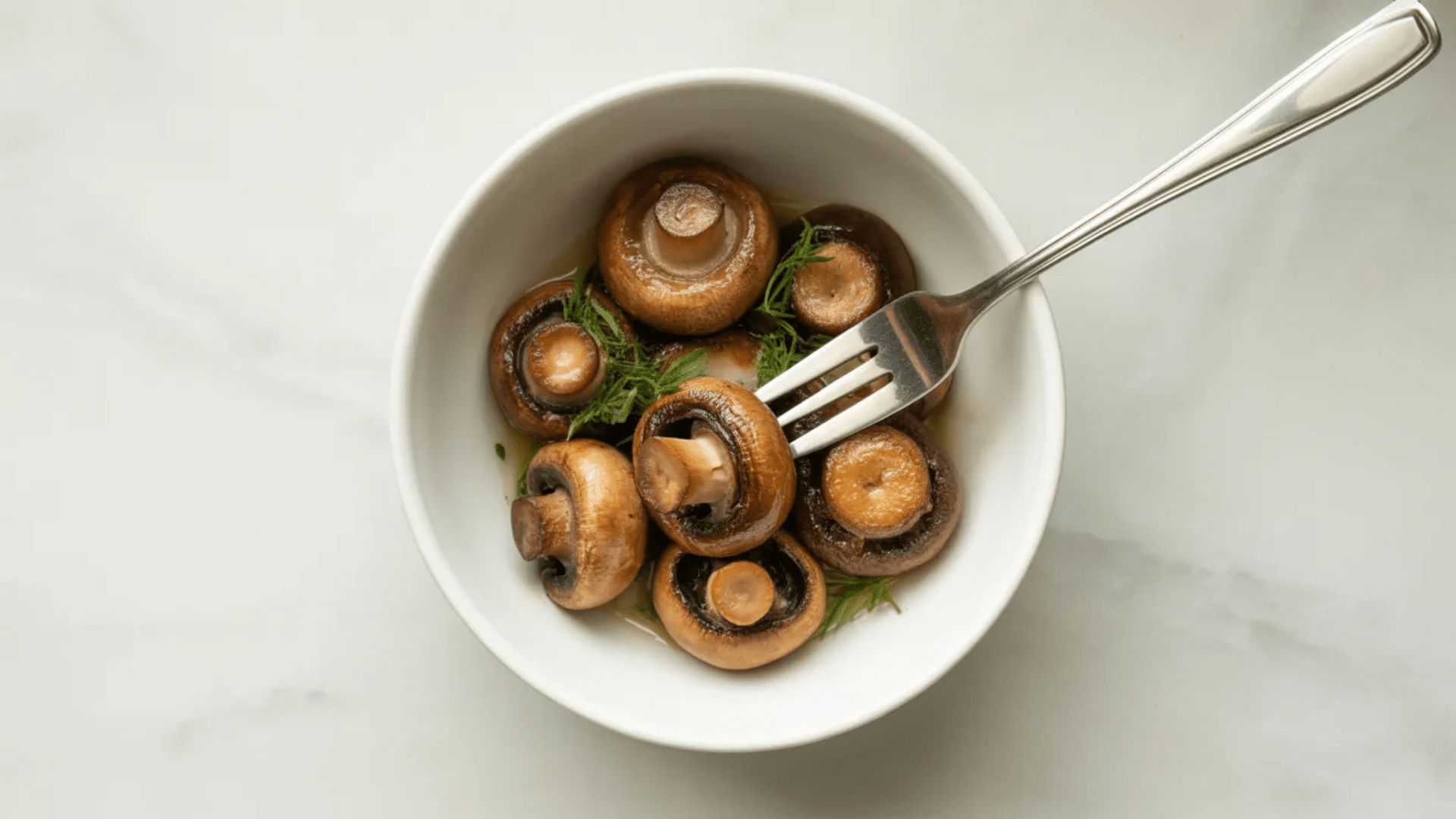 a white bowl holds several sautéed brown mushrooms garnished with fresh dill, with one mushroom being lifted by a fork
