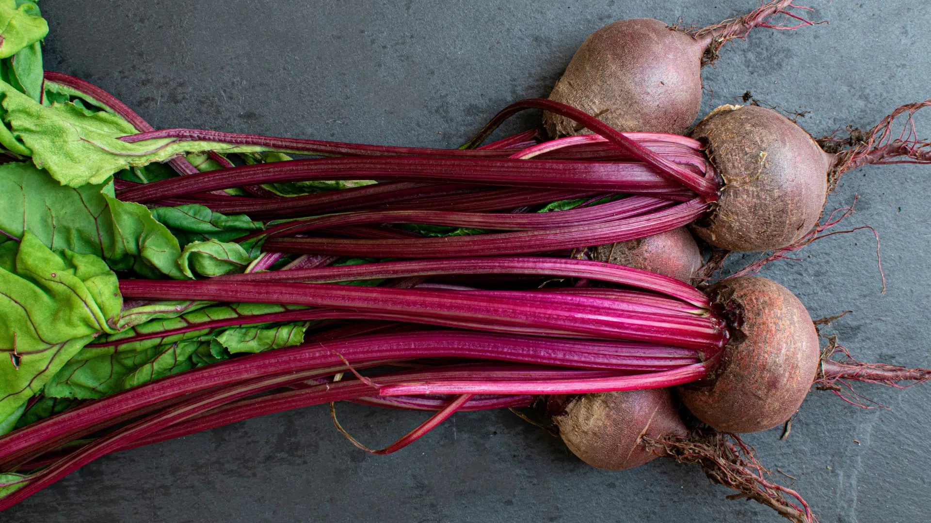 bunch of fresh beets with deep red stalks and green leaves rests horizontally against a dark, textured slate background