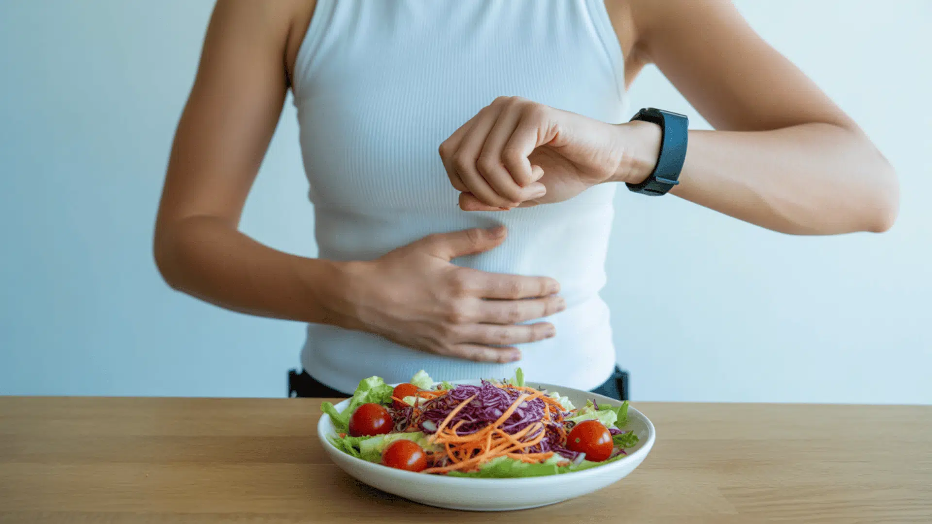 person wearing a fitness tracker holds stomach and looks at a fresh salad on a wooden table