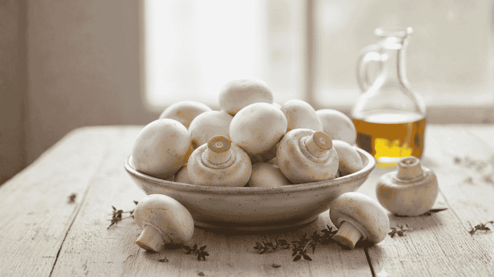 fresh white button mushrooms are piled in a ceramic bowl on a wooden table next to fresh thyme and olive oil