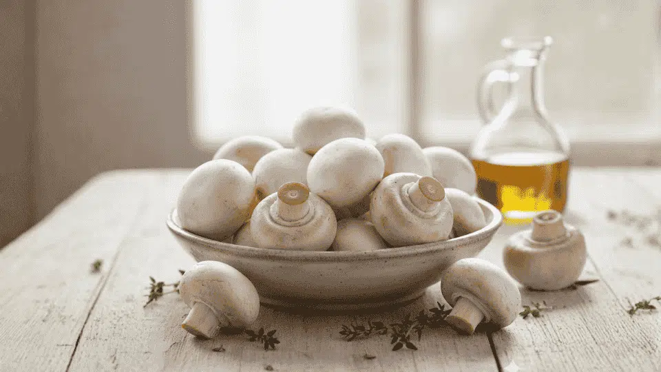 fresh white button mushrooms are piled in a ceramic bowl on a wooden table next to fresh thyme and olive oil