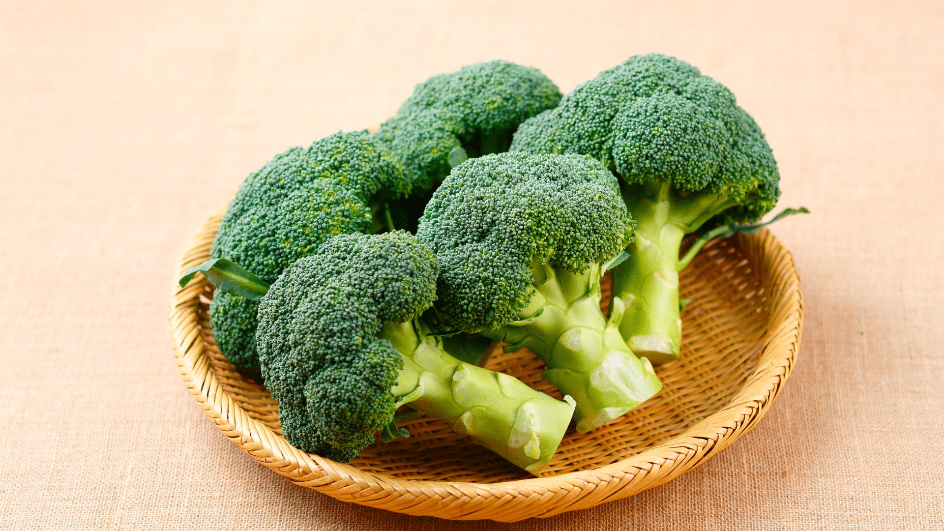 green broccoli heads with thick stalks and tightly packed florets are arranged in a woven, shallow wicker basket