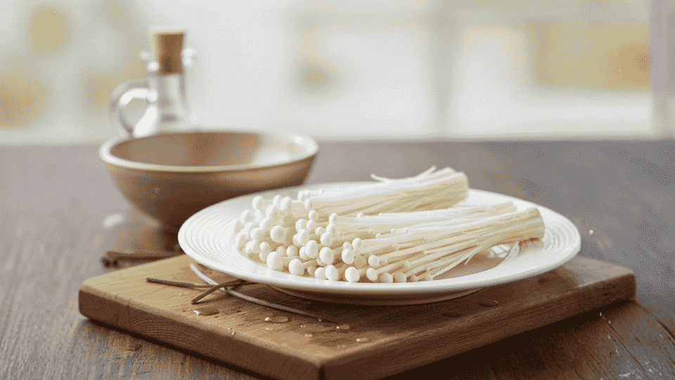 slender white enoki mushrooms with small caps are arranged on a white plate on a wooden board next to a small bowl