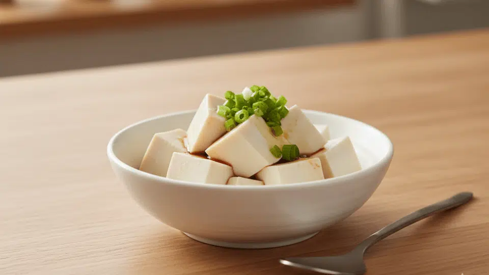 A bowl of soft silken tofu cubes topped with green onions and soy sauce on a wooden kitchen table