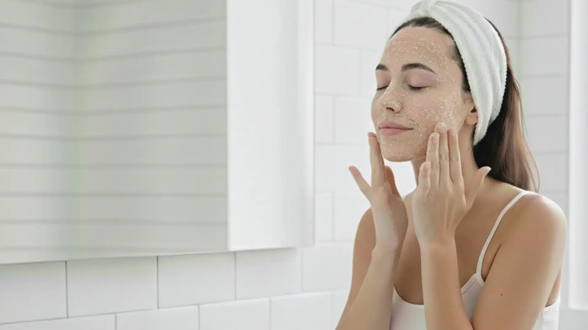 a girl applies a white exfoliating mask to her cheeks and nose in a bright bathroom, focused on her skincare routine