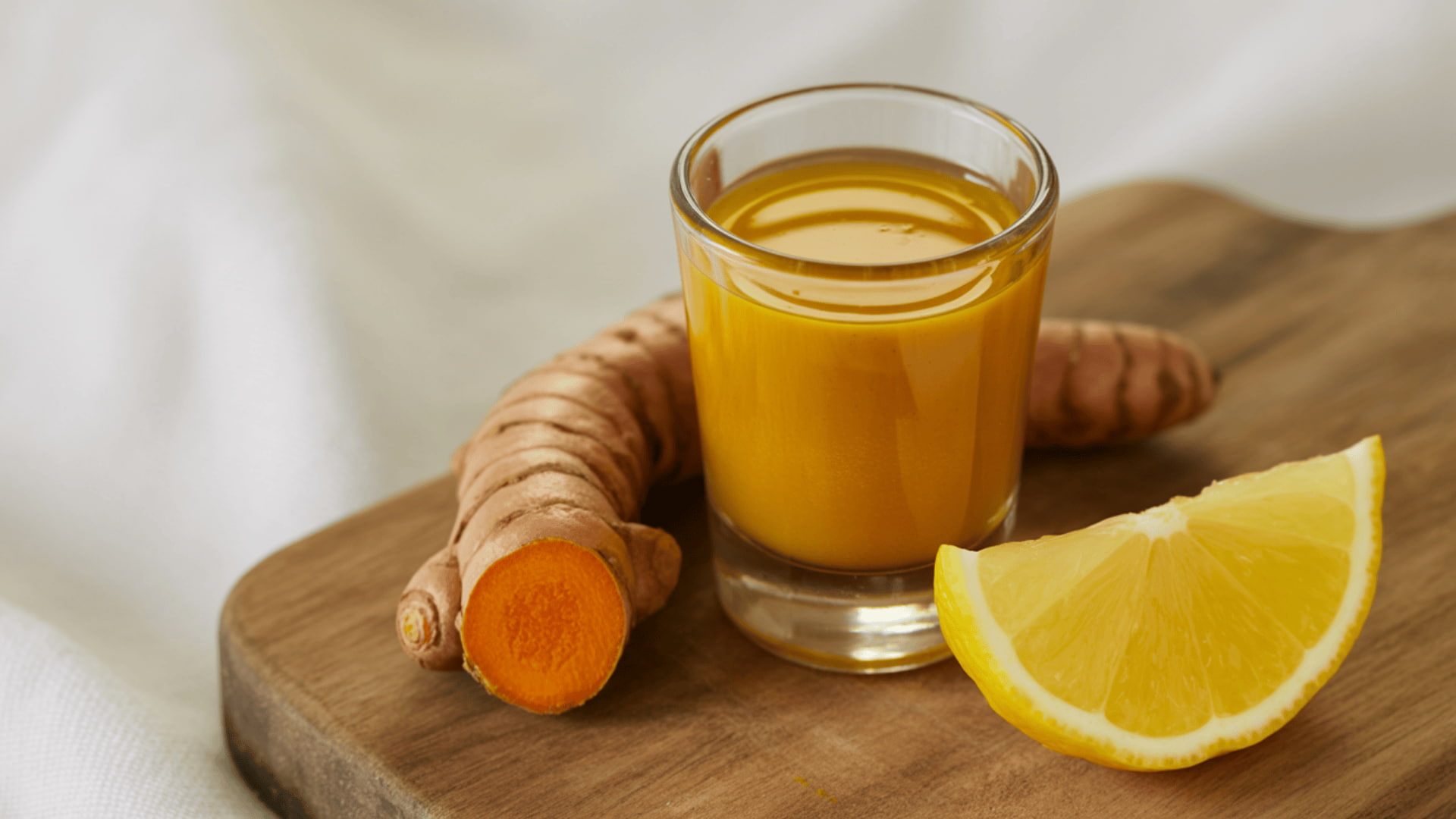 a small glass of golden turmeric shot placed next to fresh turmeric root and a lemon slice on a wooden board