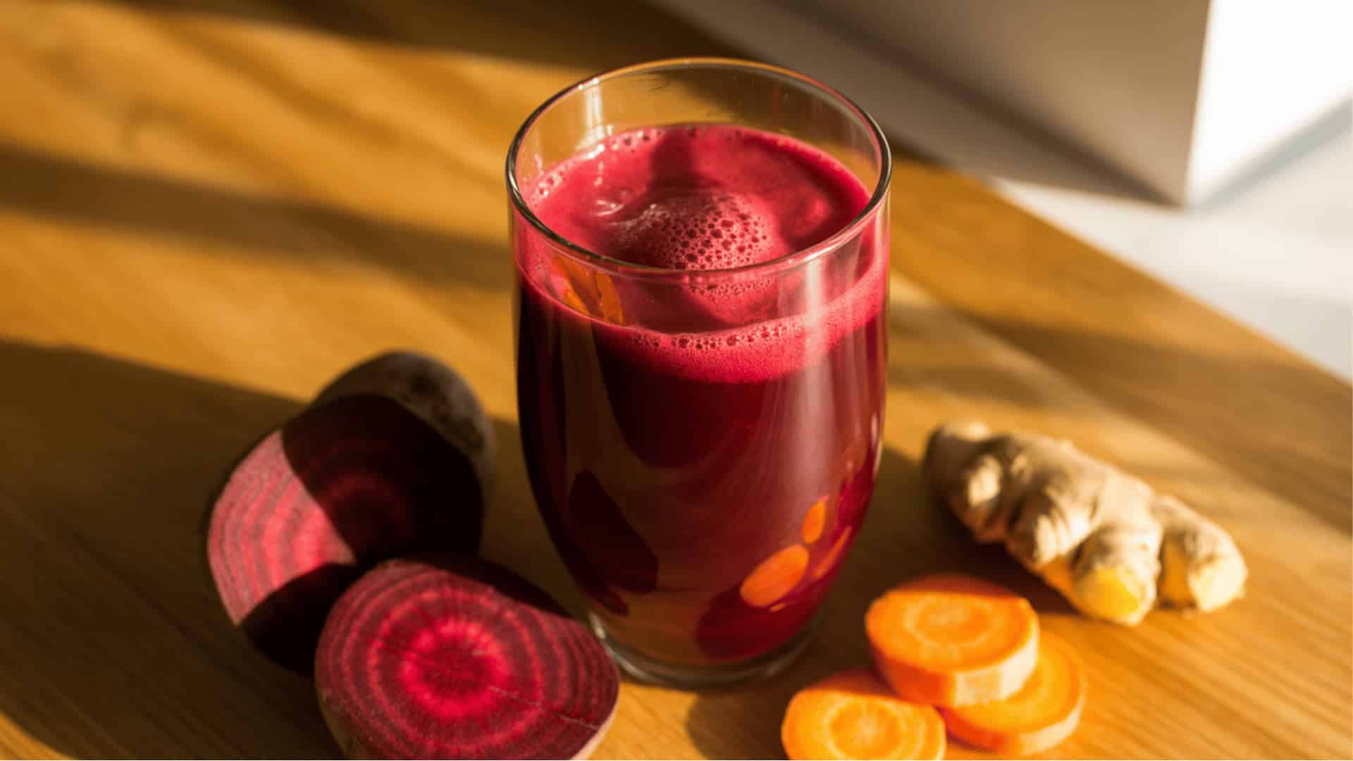 a tall glass of freshly made beetroot and carrot juice, with sliced beets, carrots, and ginger on a wooden surface