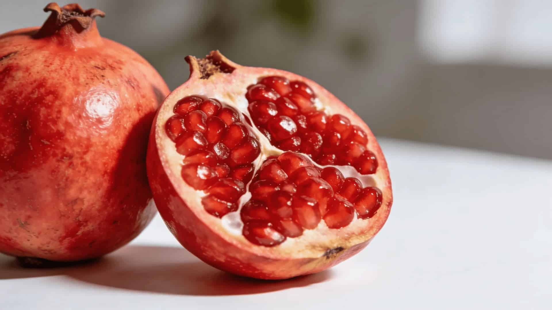 a whole pomegranate beside a halved one showing bright red seeds on a clean white surface