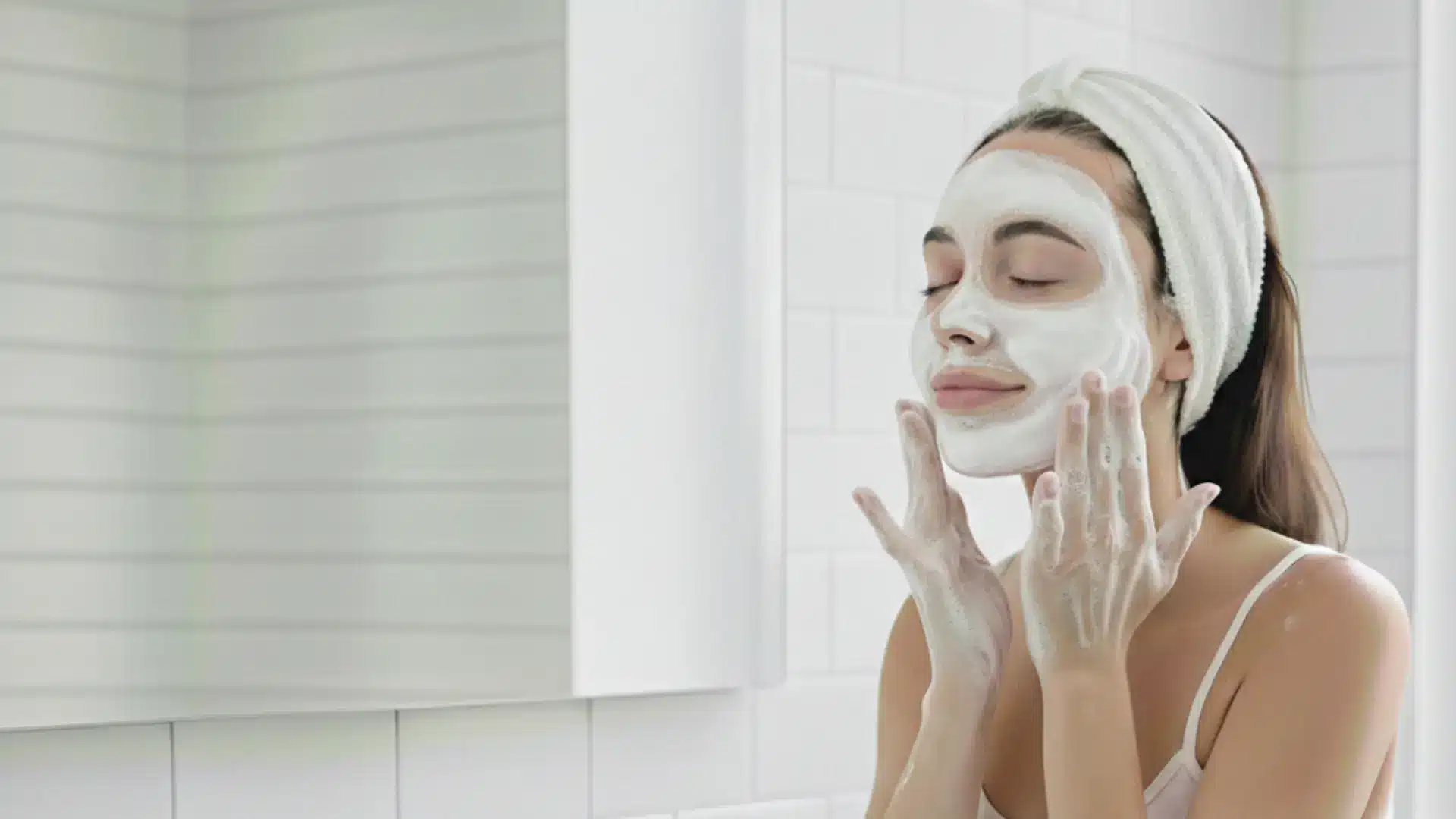 a woman in a white headband applies a foaming cleanser in a bright bathroom, promoting a fresh and clean complexion