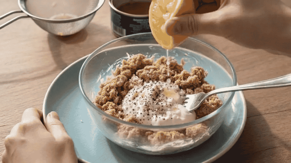 adding greek yogurt, lemon juice, and seasonings to a bowl of flaked tuna with a fork