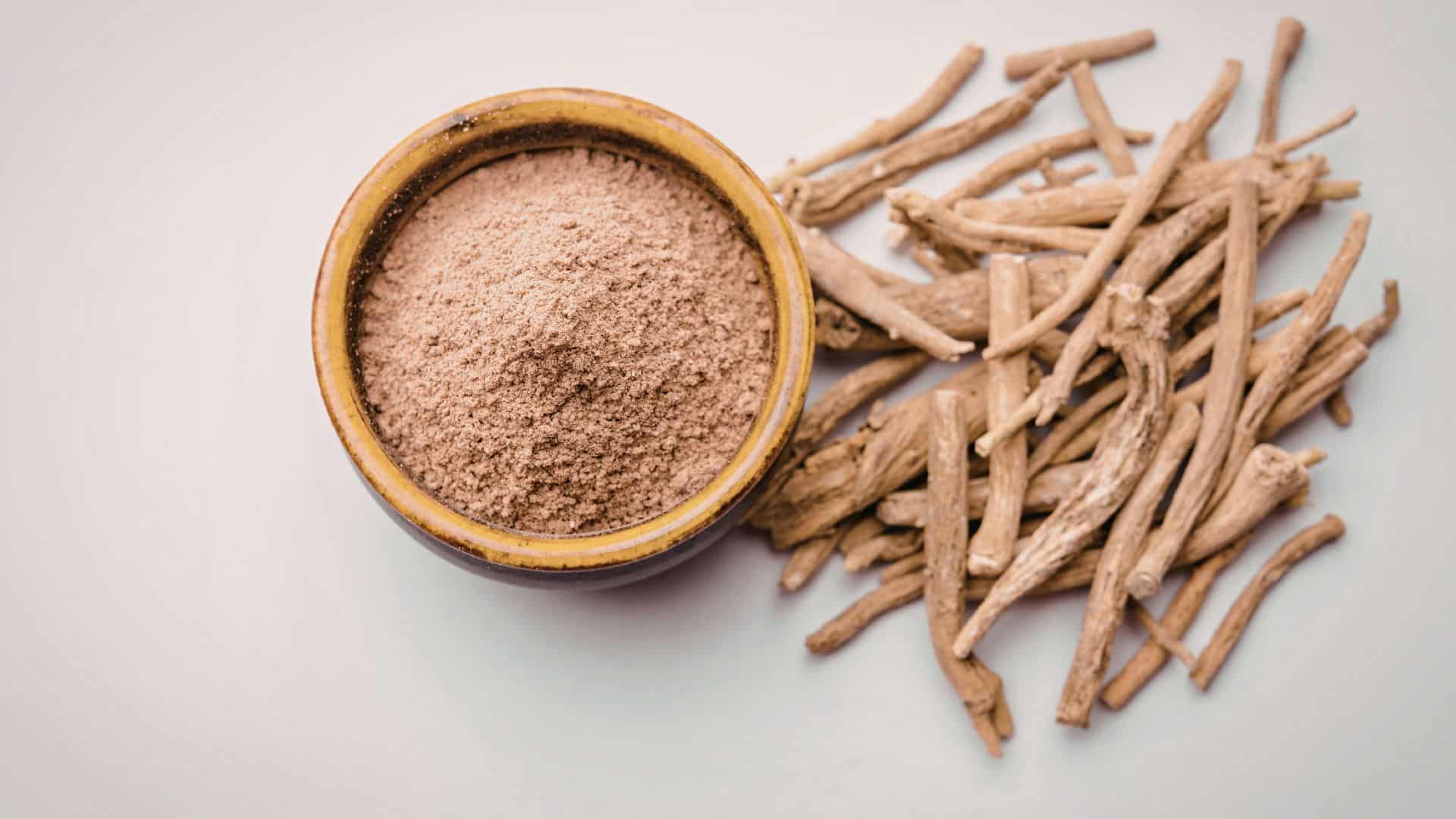 bowl of finely ground ashwagandha powder sits next to a pile of dried, raw ashwagandha roots on a white surface