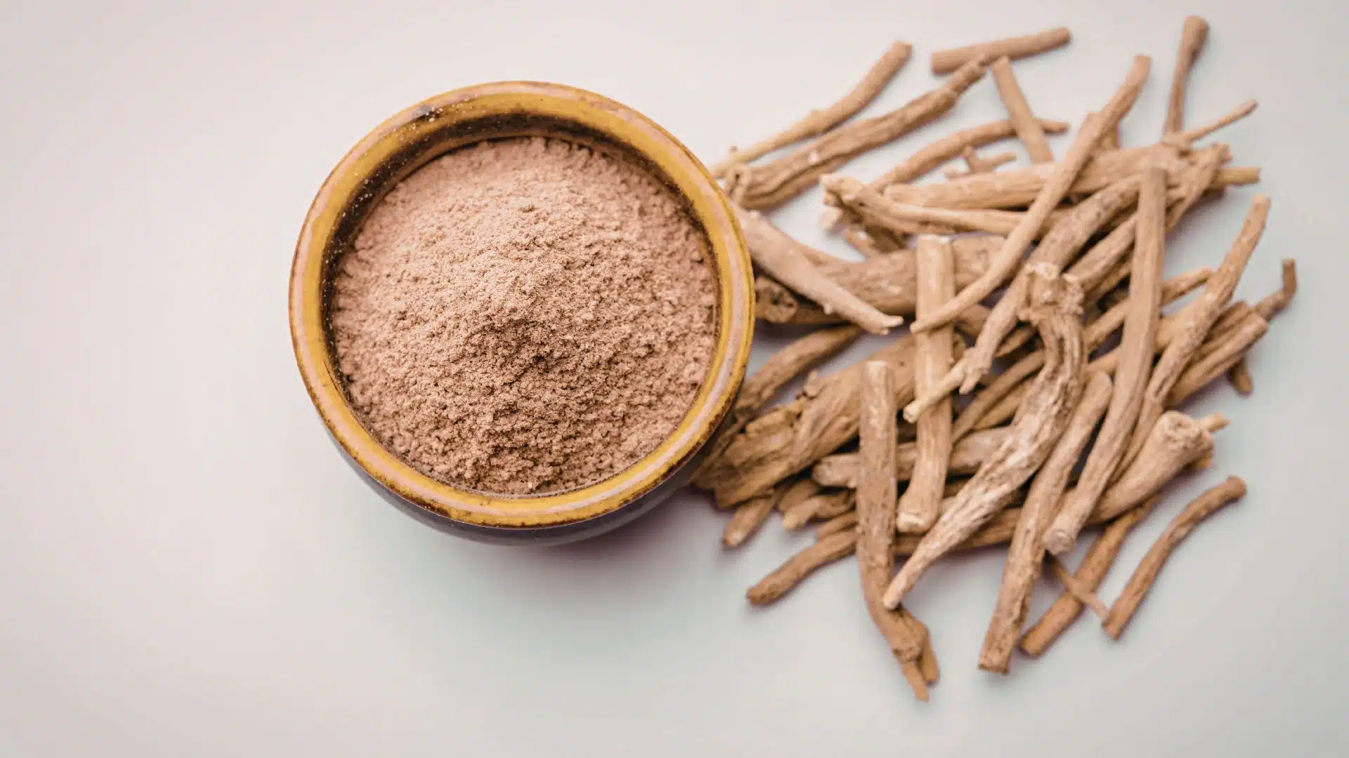 bowl of finely ground ashwagandha powder sits next to a pile of dried, raw ashwagandha roots on a white surface