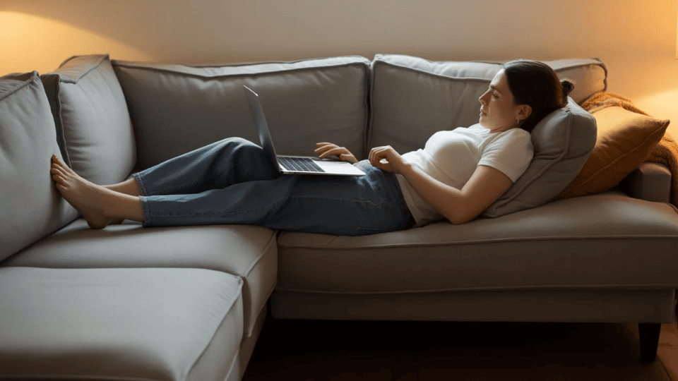 person slouching on couch while using laptop with legs stretched out showing poor posture that can strain the lower back