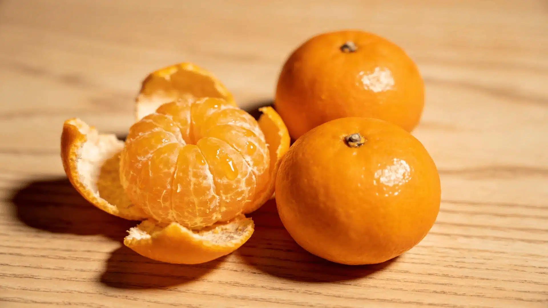 three tangerines with one peeled open arranged on a light wooden surface in soft natural lighting