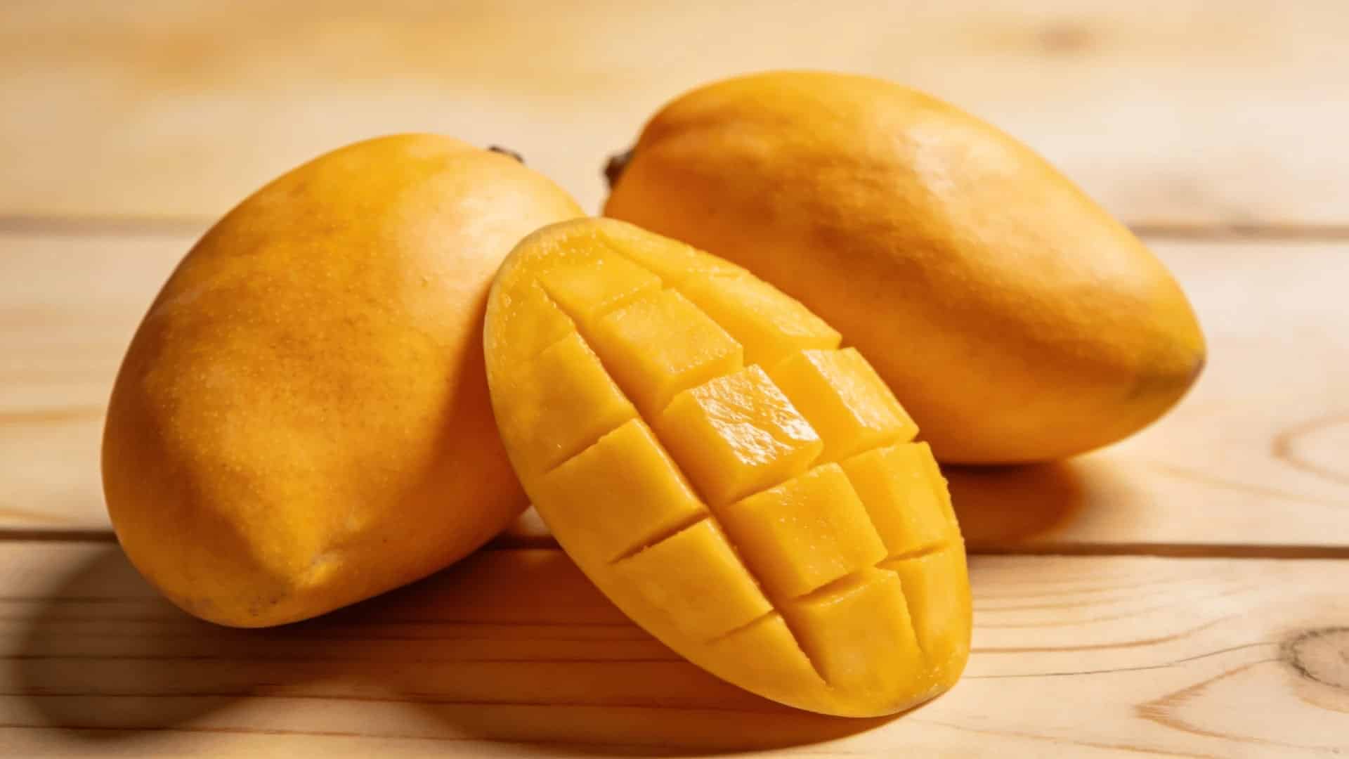 two ripe whole mangoes and one sliced mango showing yellow flesh on a light wooden surface