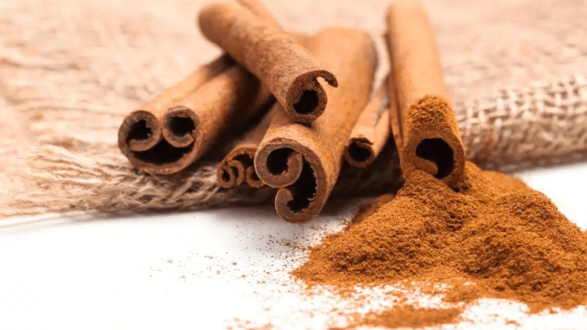 whole cinnamon sticks resting on a burlap cloth next to a pile of finely ground cinnamon powder on a white surface