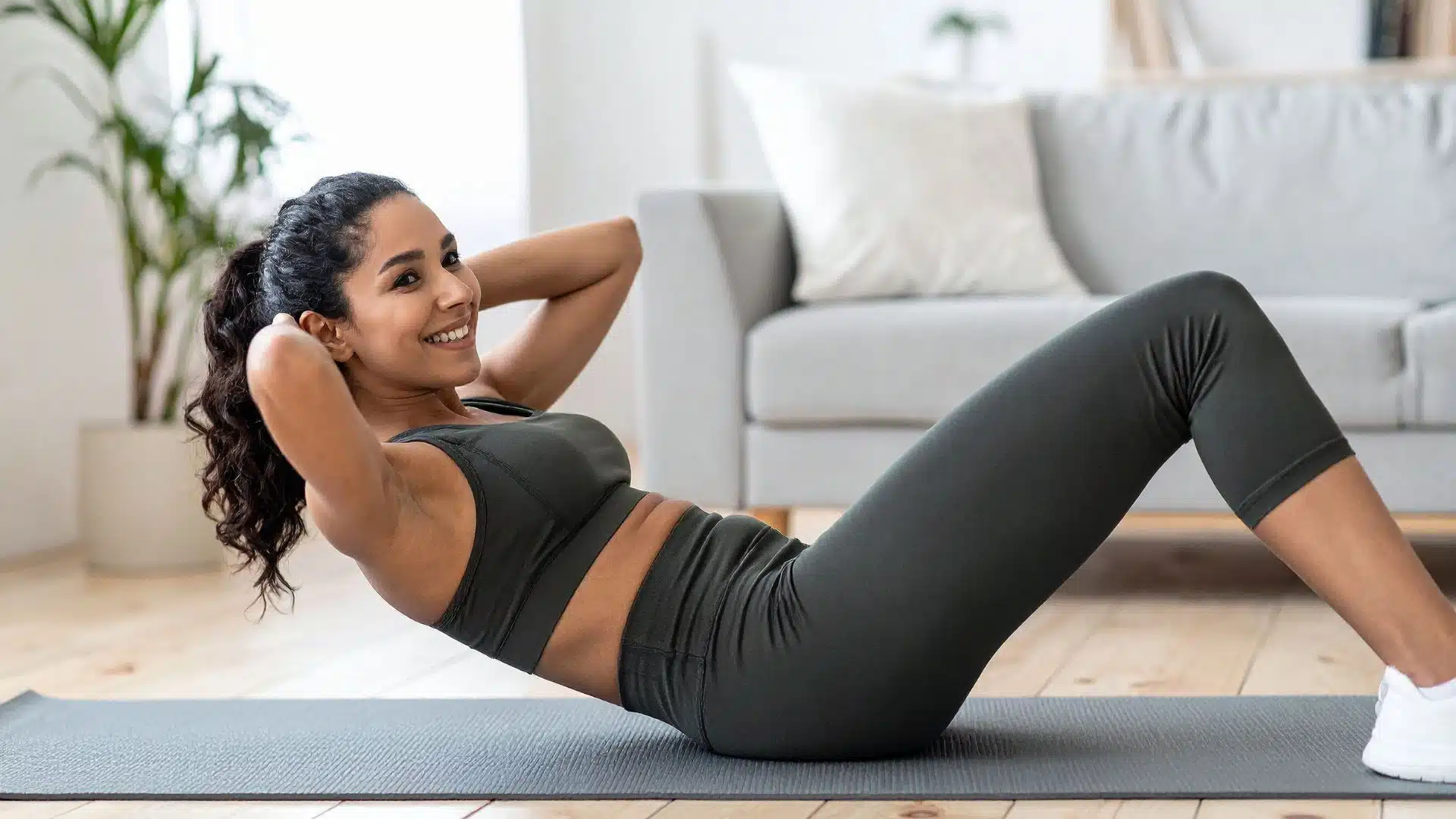 woman in dark athletic wear performing crunches on a grey yoga mat in a bright, modern living room setting