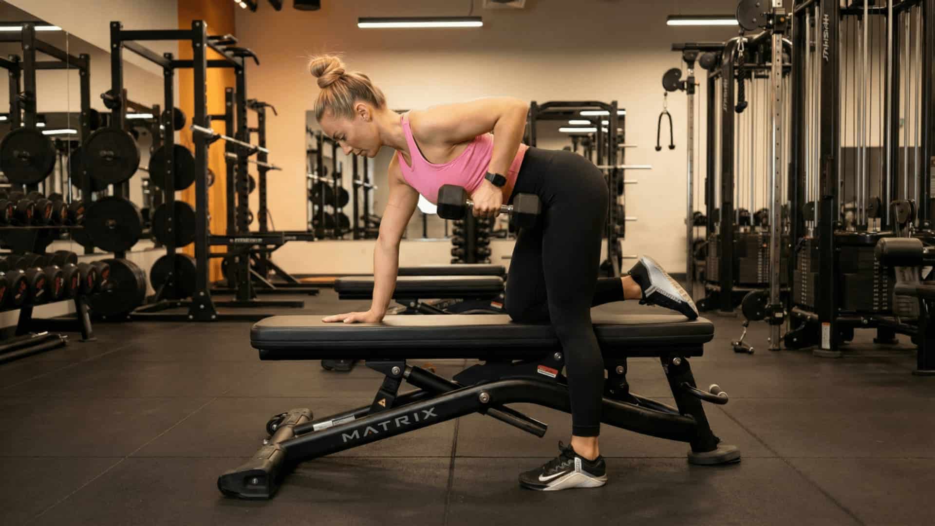 woman performing a single arm dumbbell row on a bench in a well-equipped gym