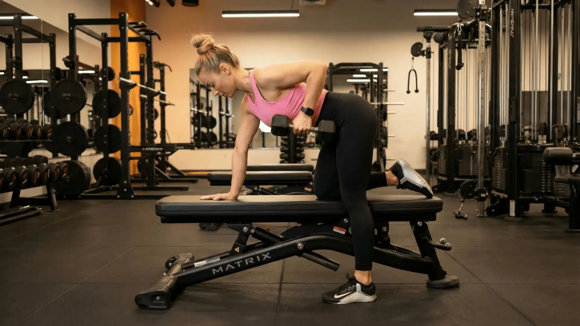 woman performing a single arm dumbbell row on a bench in a well-equipped gym