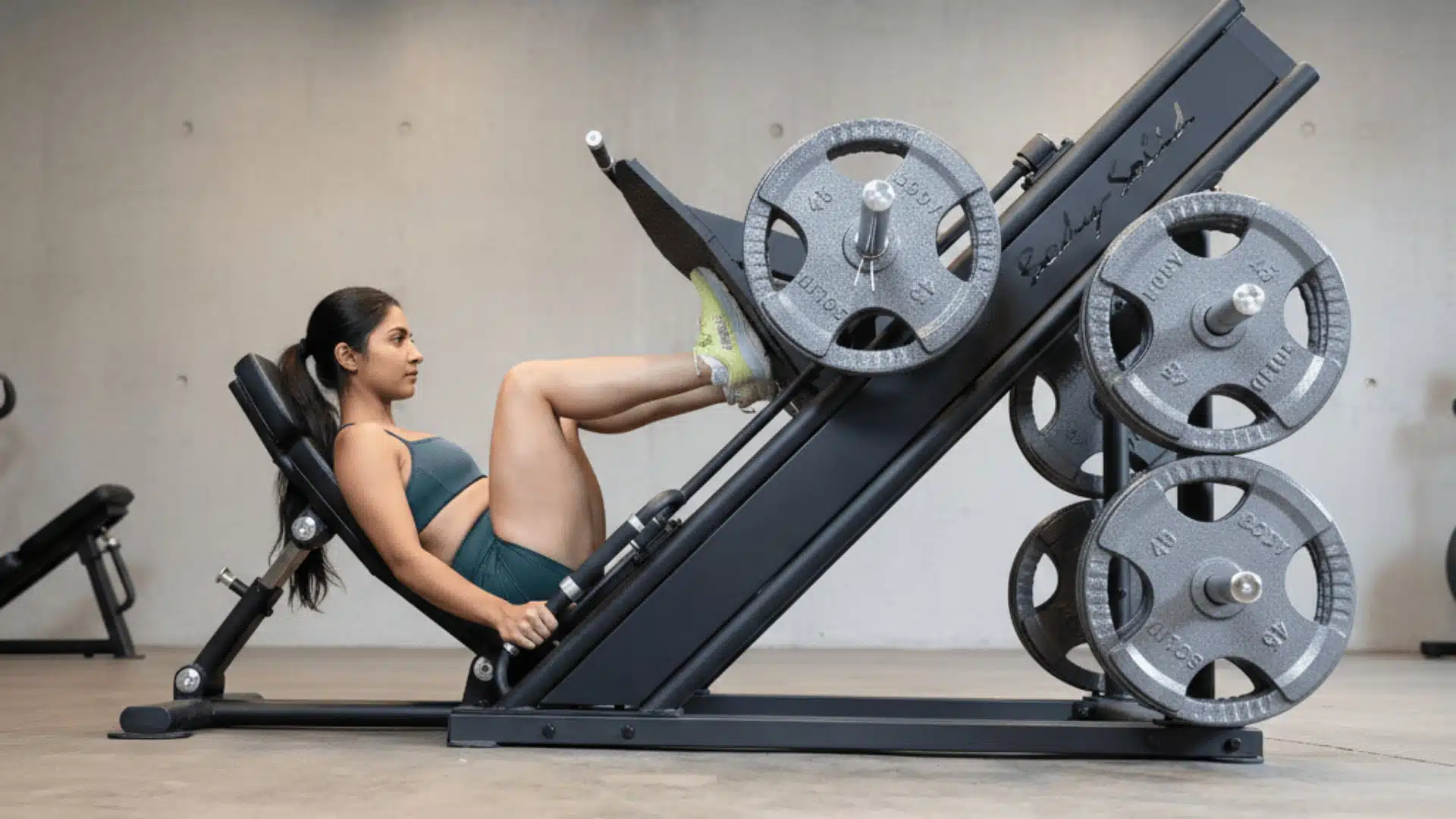 woman performing leg press on angled machine loaded with weight plates in gym setting with concrete wall background
