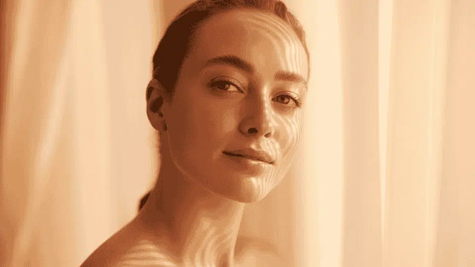 woman with hair pulled back in warm golden sunlight with light patterns on her face and shoulder looking at camera