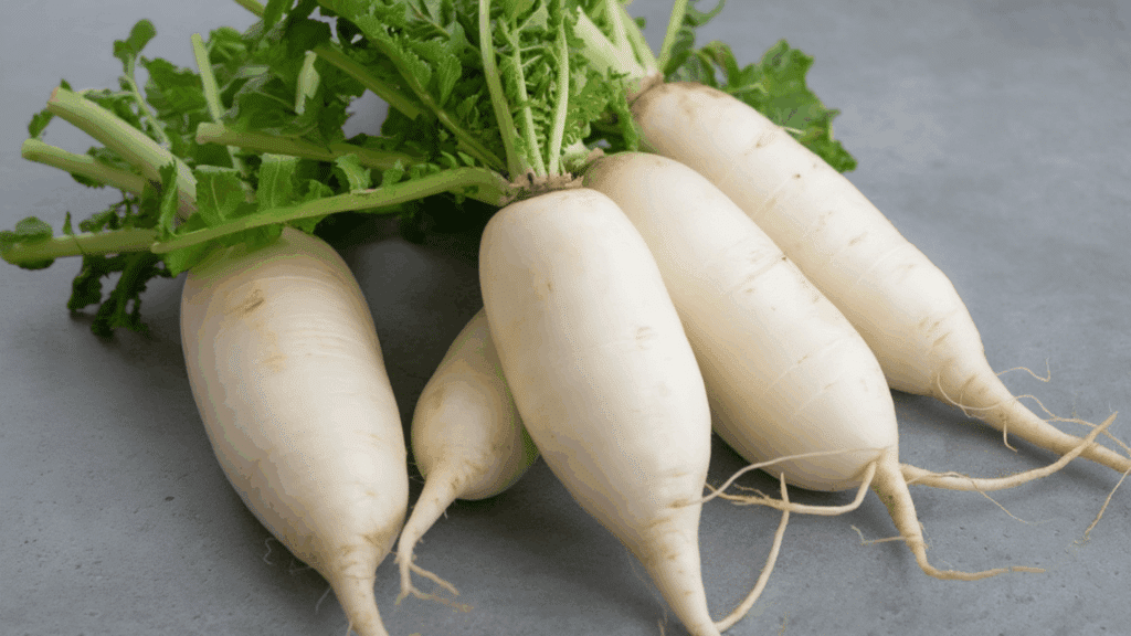 a close-up image of several daikon radishes with green tops, resting on a gray surface, showcasing their long, white roots