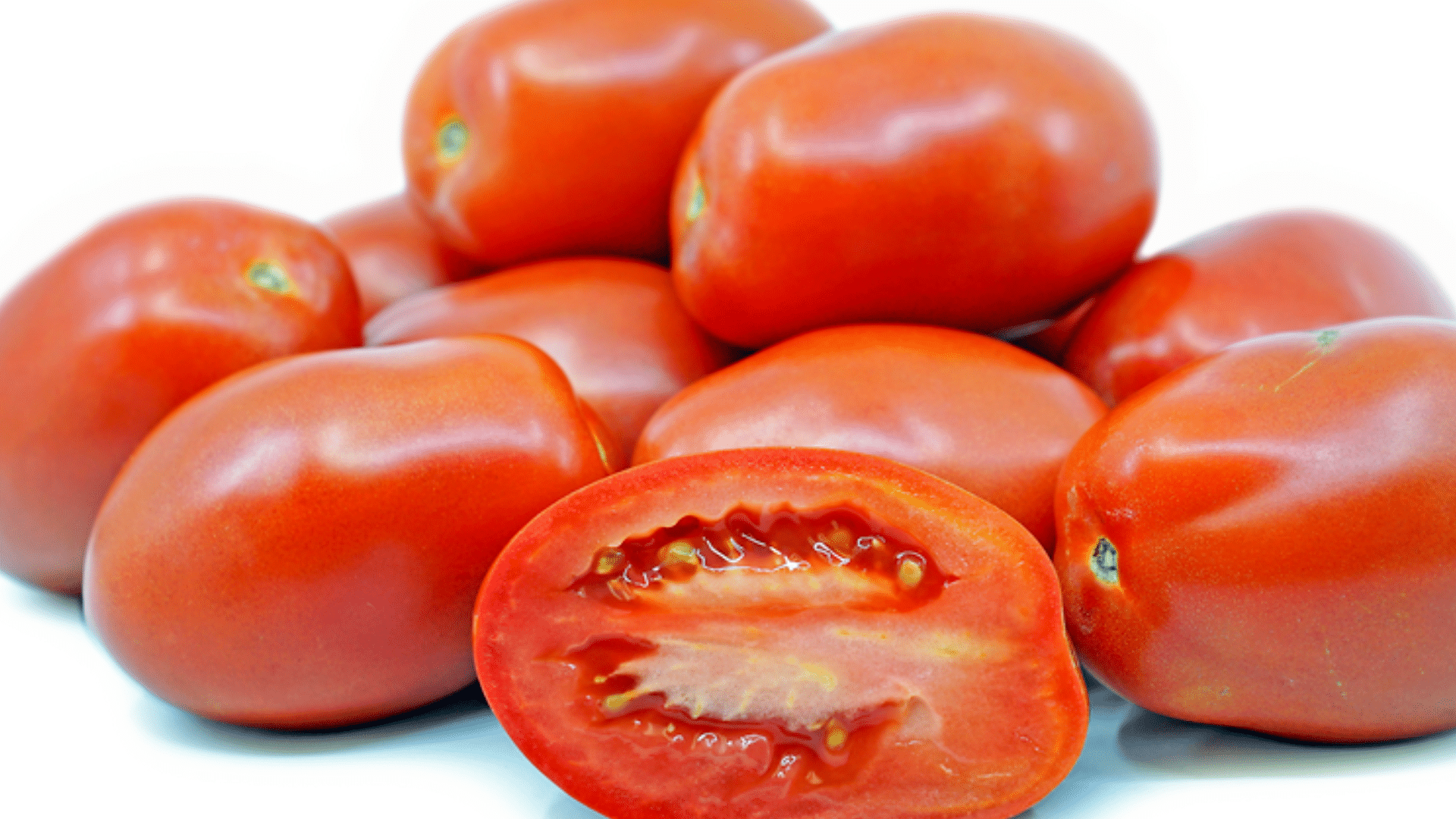 a halved roma tomato showing meaty texture and seeds on a wooden board with basil, salt, and peppers nearby