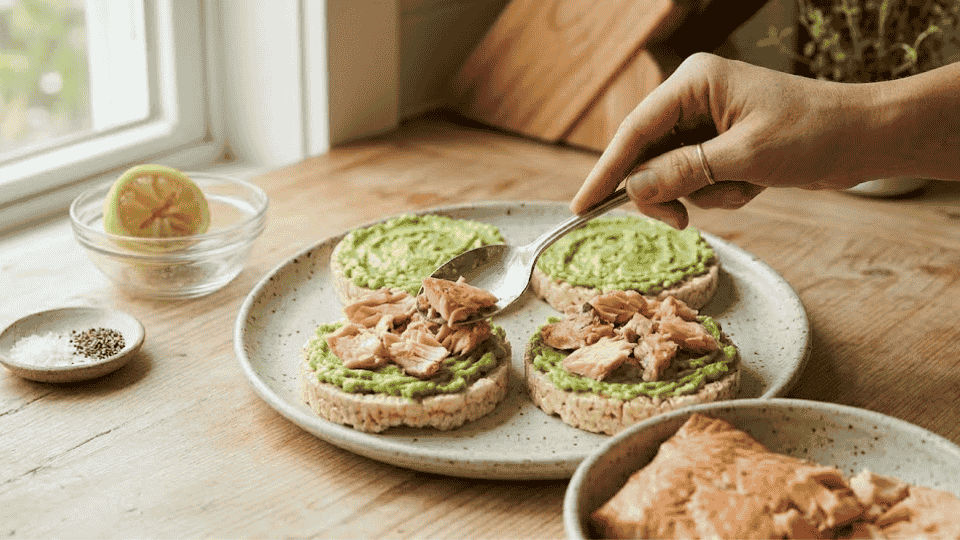 a hand adding flakes of cooked salmon over the avocado layer on rice cakes to finish the dish