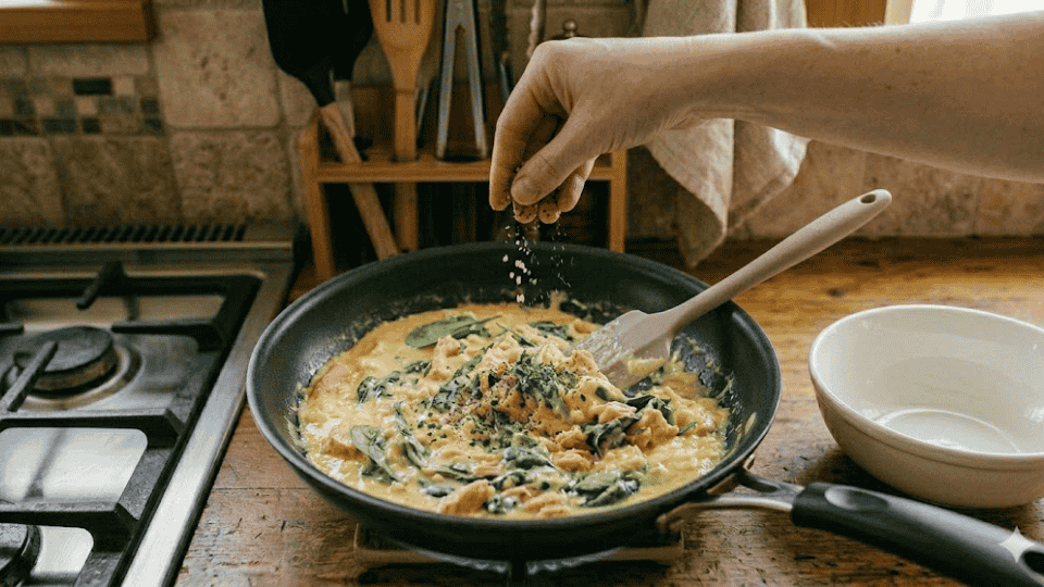 a hand sprinkles salt and pepper over the finished salmon and spinach scramble in the pan