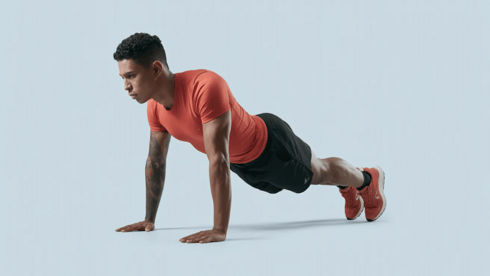 a tattooed man in a red shirt performing a push up against a light blue background