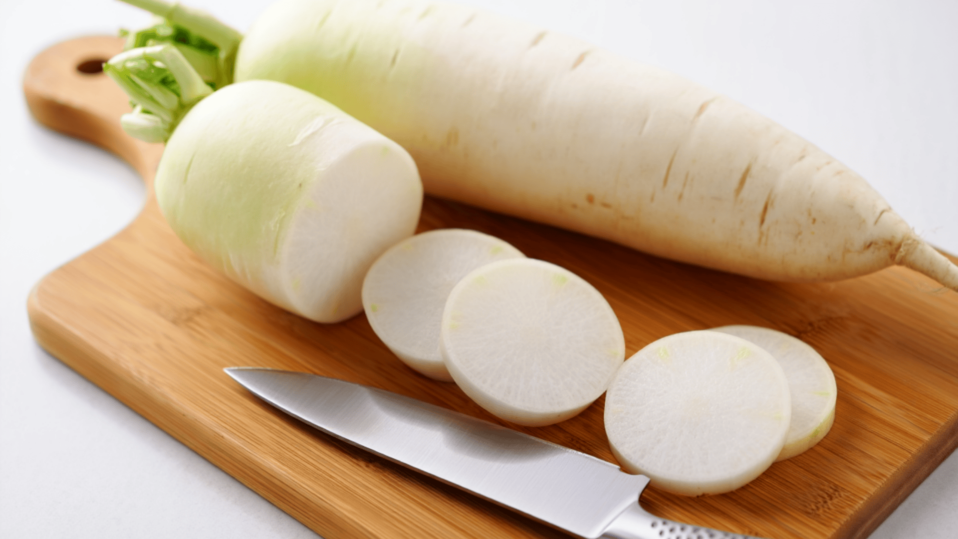 a whole daikon radish, one large cut piece, and three slices rest on a wooden board next to a sharp chef knife