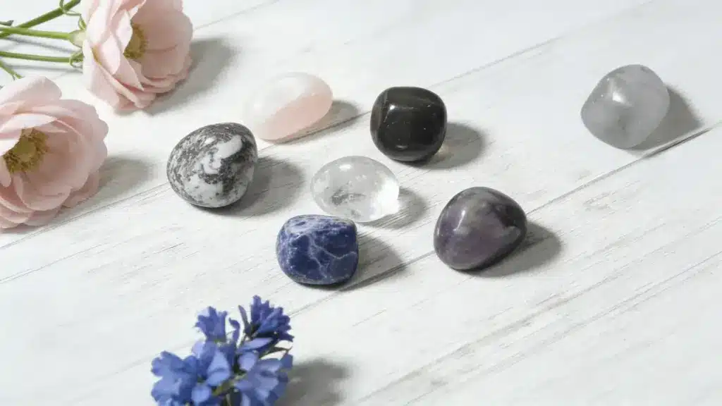 assortment of tumbled healing crystals and soft pink and blue flowers on a light-colored wooden background