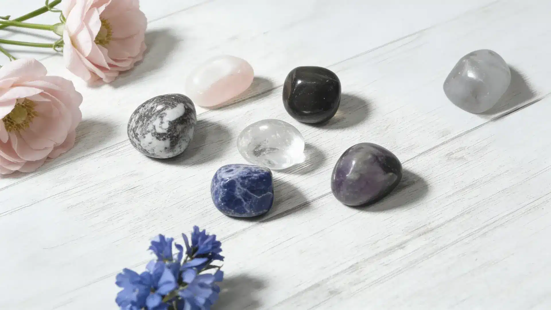 assortment of tumbled healing crystals and soft pink and blue flowers on a light-colored wooden background