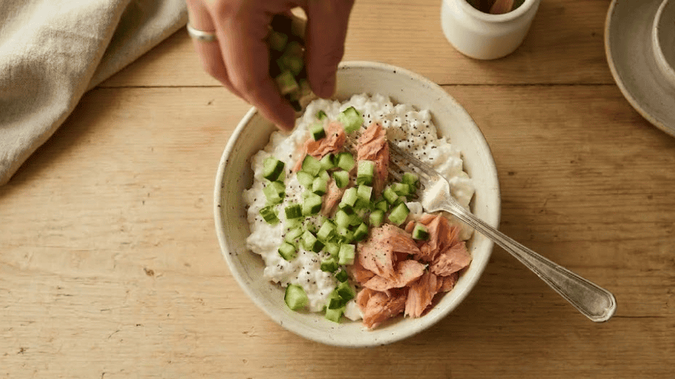 chopped cucumber added to a cottage cheese and salmon bowl on a light wooden kitchen counter