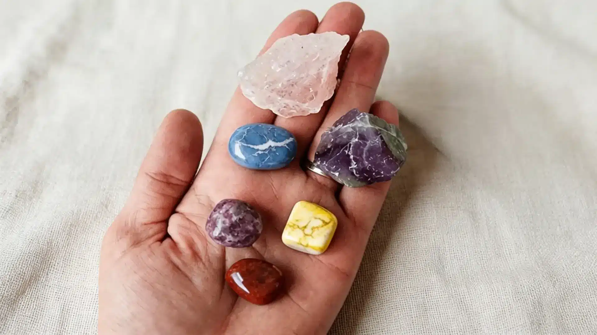 close-up of a person's hand holding a single raw amethyst crystal against a soft neutral fabric background