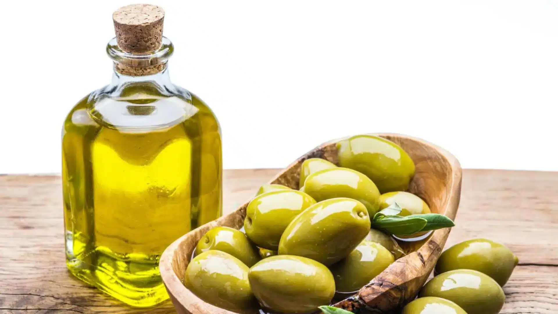 corked glass bottle of golden olive oil rests on a wooden surface beside a rustic bowl of green olives