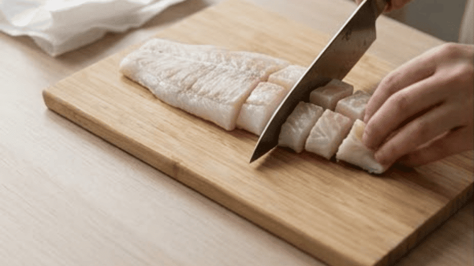 cutting cod fillet into small pieces on wooden board in soft natural light