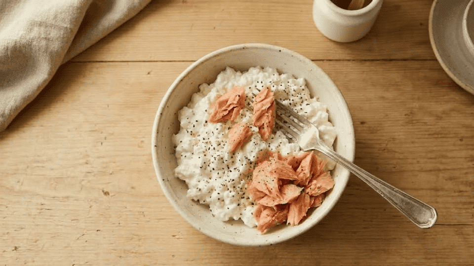 flaked salmon being added to cottage cheese in a bowl on a light wooden kitchen counter