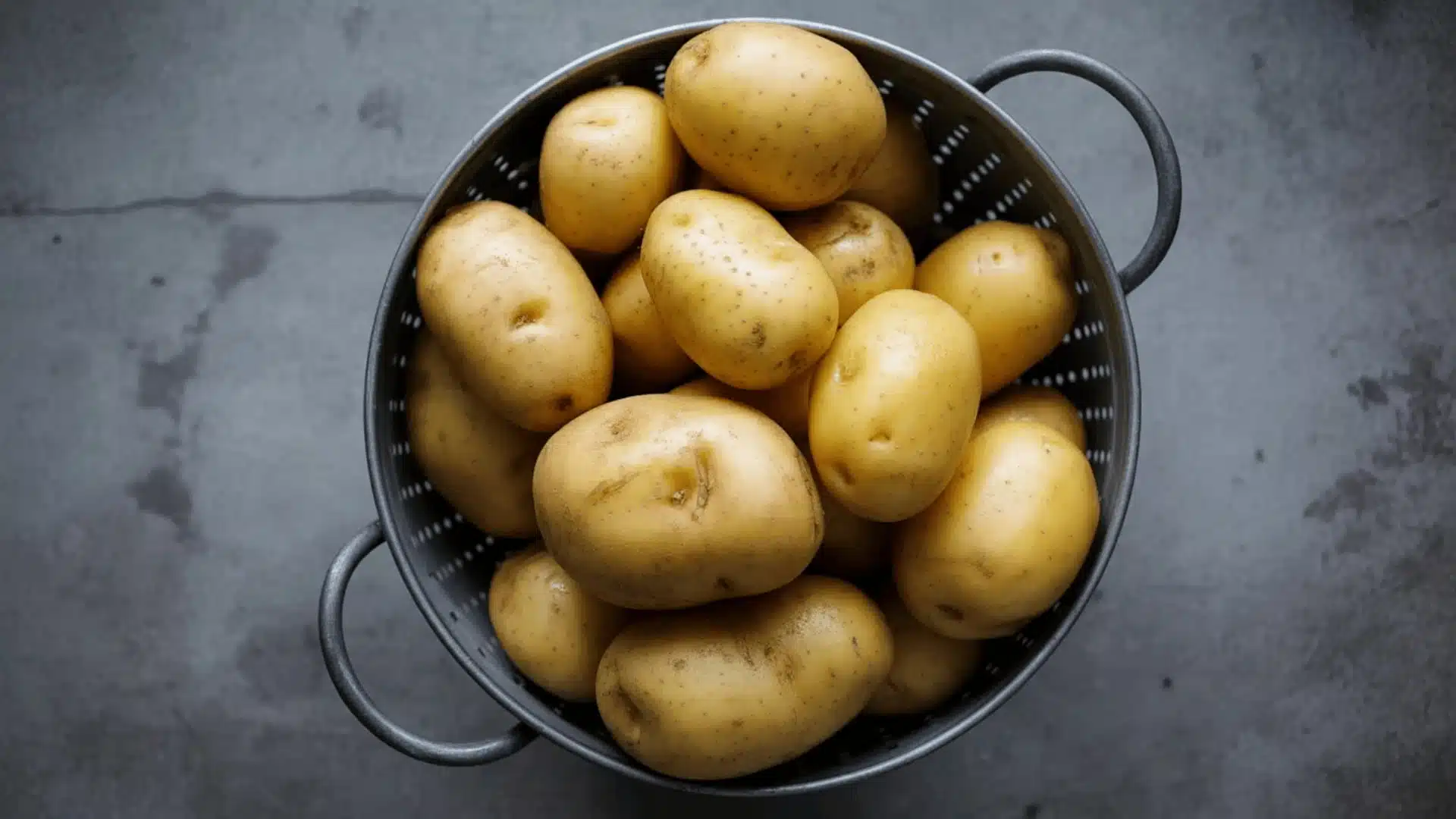 fresh yellow yukon gold potatoes piled in a grey perforated metal colander on a grey concrete surface