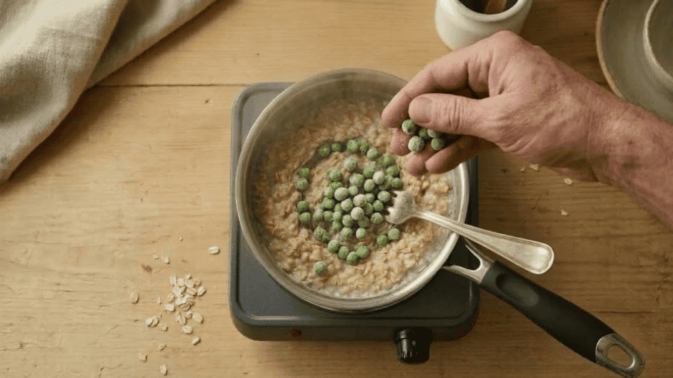 frozen peas added to a saucepan of cooking oatmeal on a light wooden kitchen counter