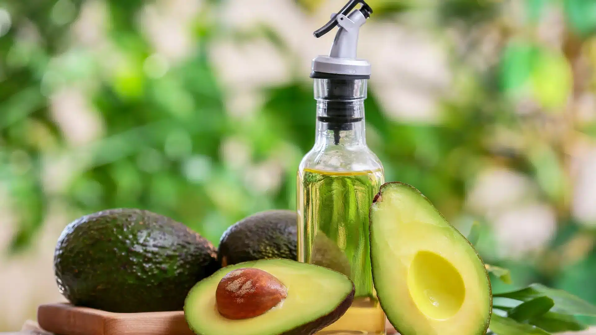 glass bottle of avocado oil stands behind several halved and whole ripe avocados on a rustic wooden table setup