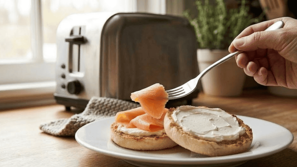 hand using a fork to layer smoked salmon slices onto a cream cheese covered english muffin