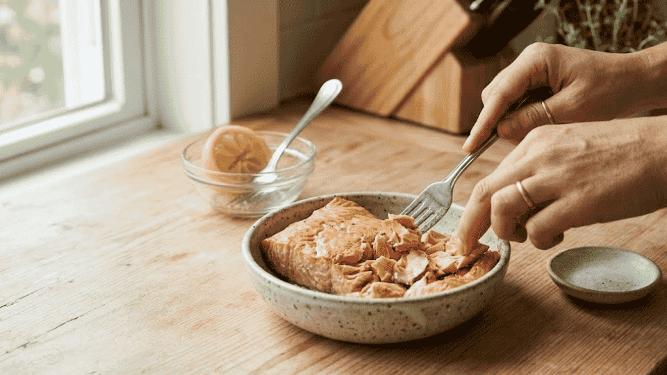 hands flaking a cooked salmon fillet with a fork in a bowl on a rustic wooden kitchen surface