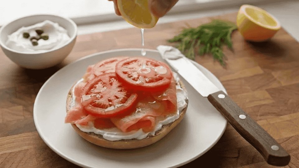 squeezing lemon a single bagel half on a white plate topped with cream cheese, smoked salmon, tomato, and capers