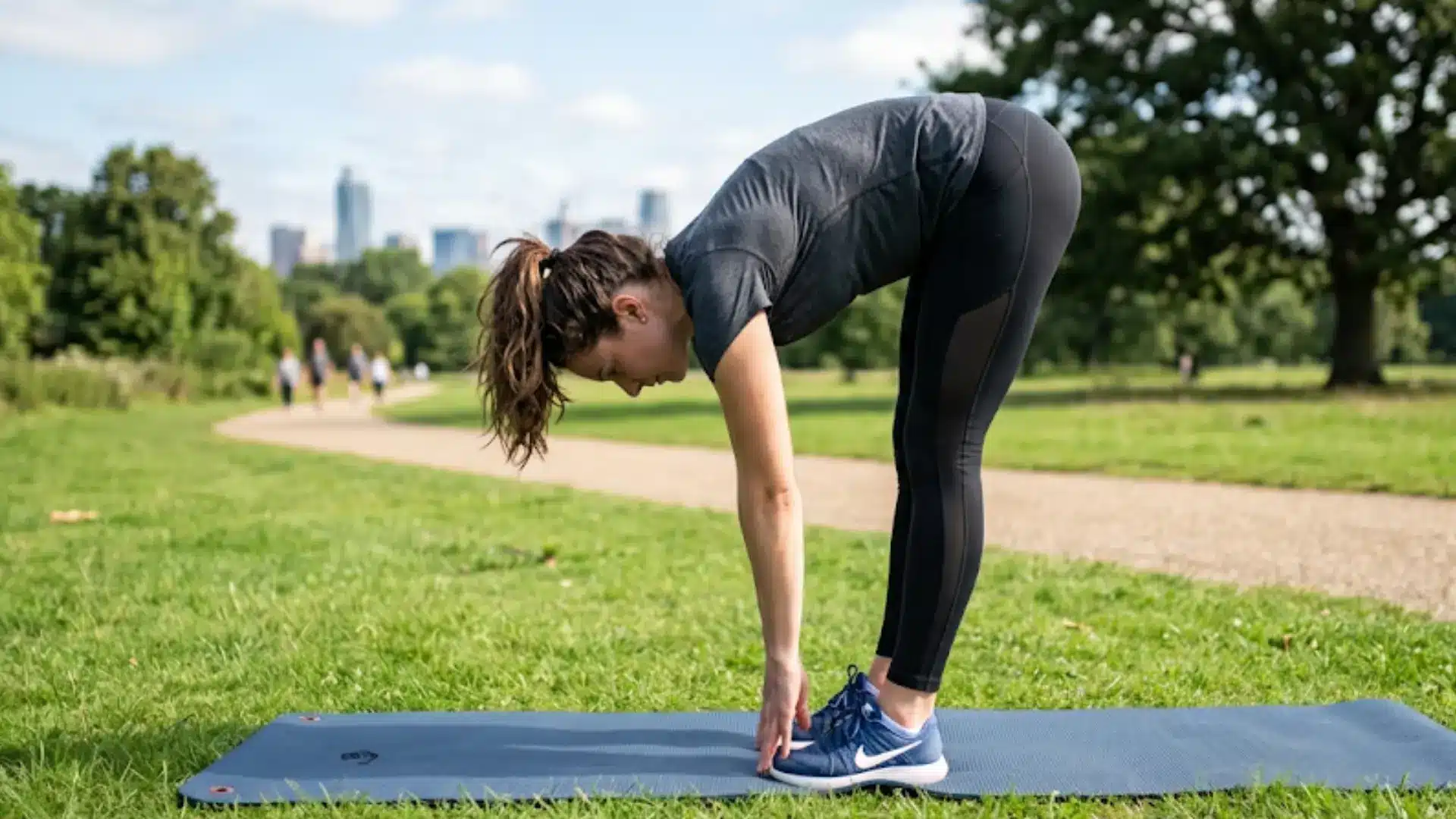 woman in fitness gear performs standing toe touches on a blue mat in a sunny park with city views